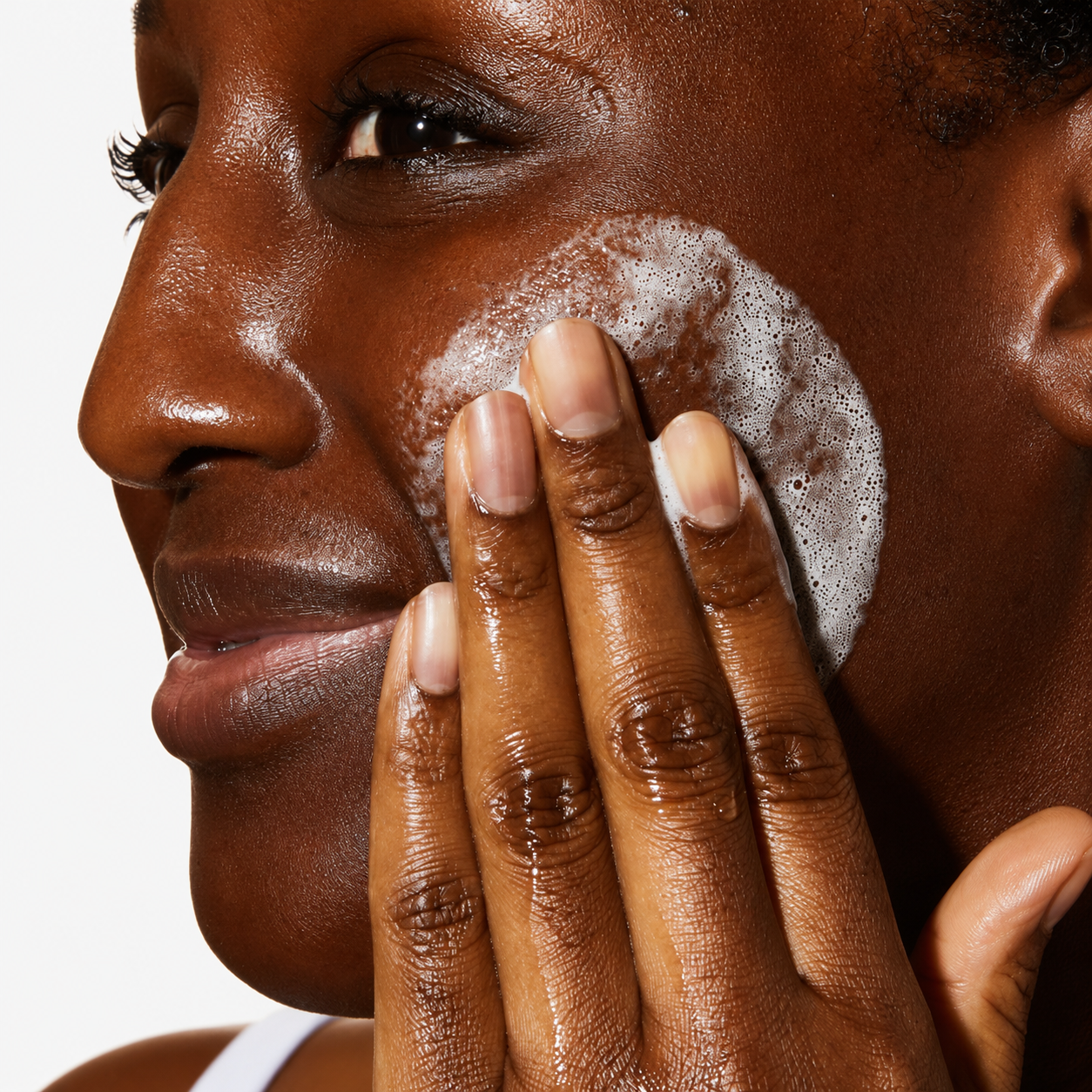 Close-up of a person applying a white foaming cleanser or scrub to their cheek with their fingers, shown in profile against a light gray background.