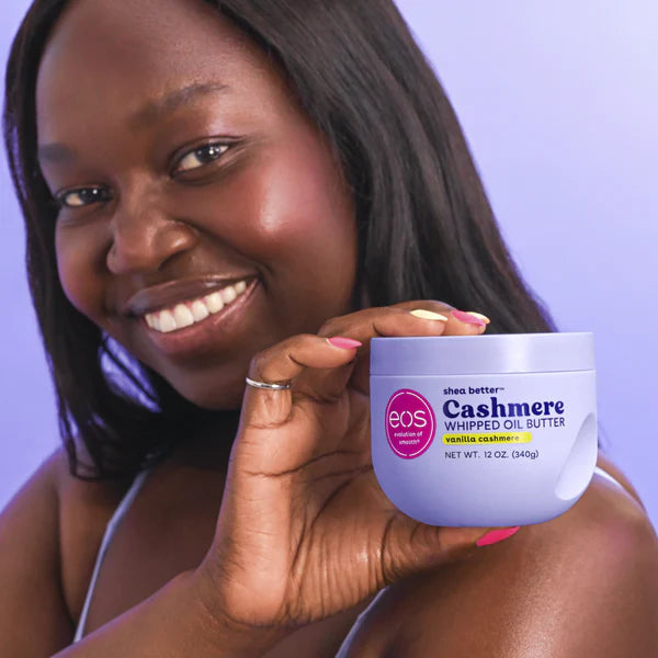 A smiling woman holds up a white jar of EOS Cashmere Whipped Oil Butter against a light purple background.